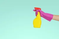 Hand in purple glove holding yellow spray bottle against blue background.